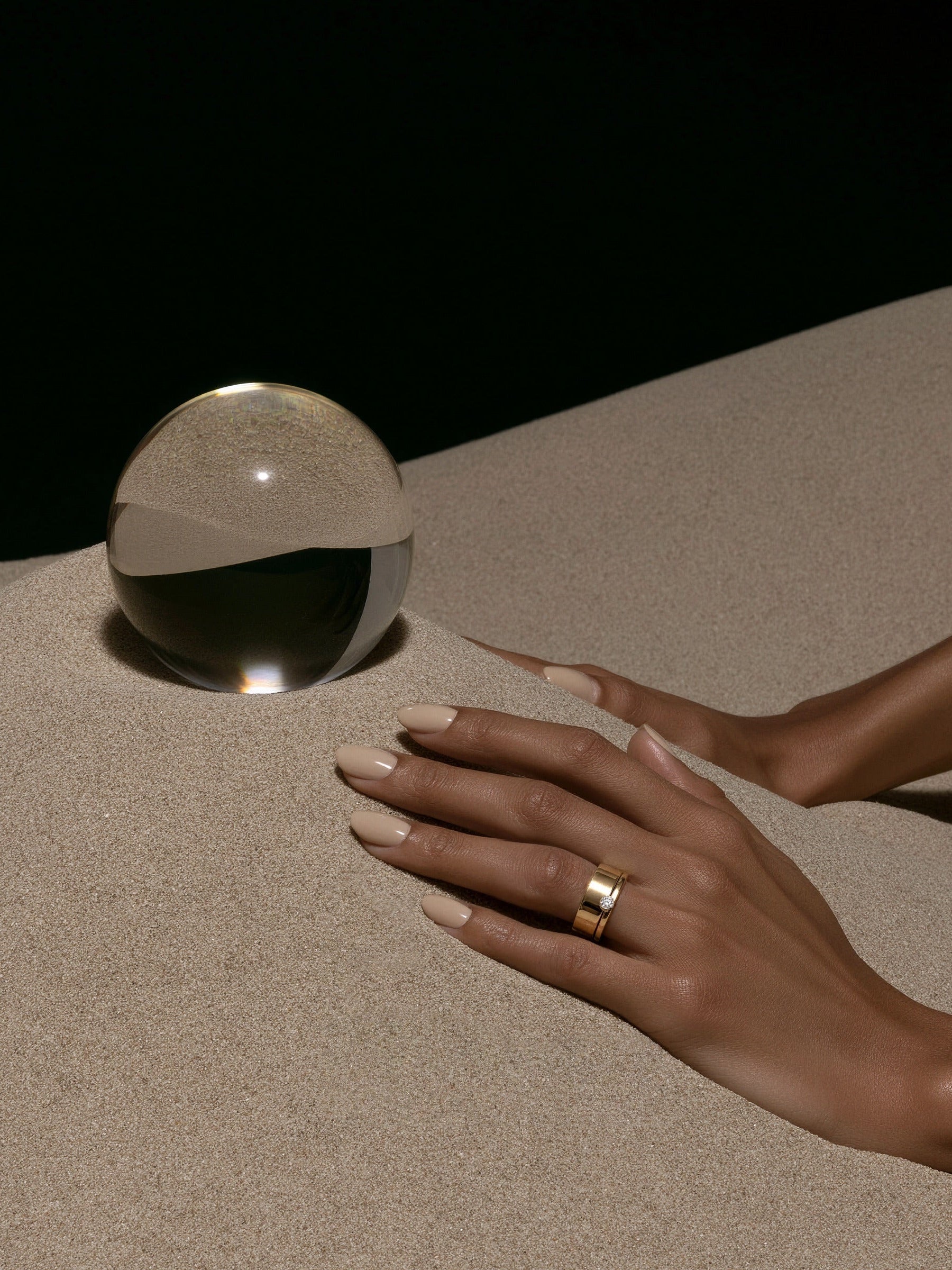 Hand mit MIONA Aufsteckring in Gold neben einer Glaskugel im Sand