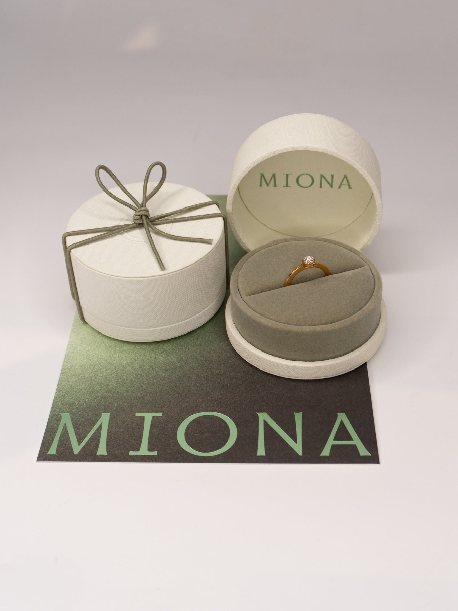 MIONA Ring in der MIONA Schmuckdose, daneben die verschlossene Dose mit Schleife auf der Markenkarte